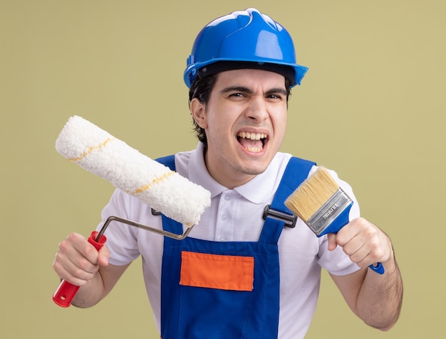 Young builder man in construction uniform and safety helmet holding paint roller and brush shouting with aggressive expression standing over green wall