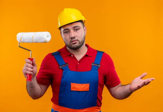 Young builder man in construction uniform and safety helmet holding paint roller looking confused spreading arms to the sides 