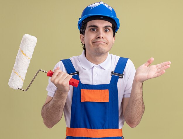 Young builder man in construction uniform and safety helmet holding paint roller looking at front confused shrugging shoulders standing over green wall