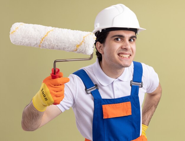 Young builder man in construction uniform and safety helmet holding paint roller looking at front happy and cheerful smiling standing over green wall