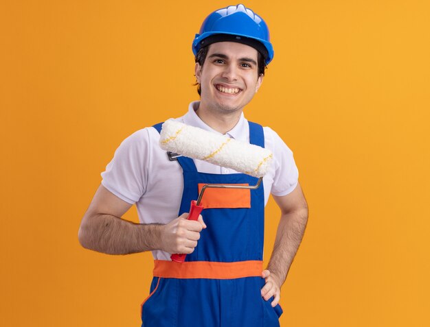 Молодой строитель в строительной форме и защитном шлеме, держащий валик с краской, смотрит вперед с улыбкой на лице, стоя над оранжевой стеной