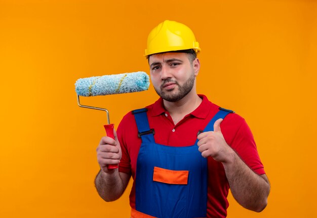 Молодой строитель в строительной форме и защитном шлеме, держащий валик с краской, показывает палец вверх