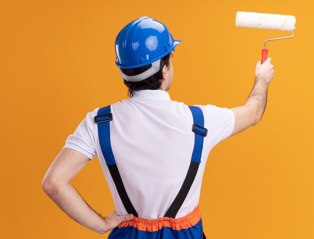 Young builder man in construction uniform and safety helmet holding paint roller standing with his back over orange wall