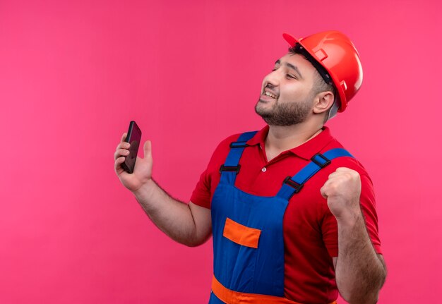 Молодой строитель в строительной форме и защитном шлеме держит смартфон, сжимая кулак, счастлив и выходит, радуясь своему успеху