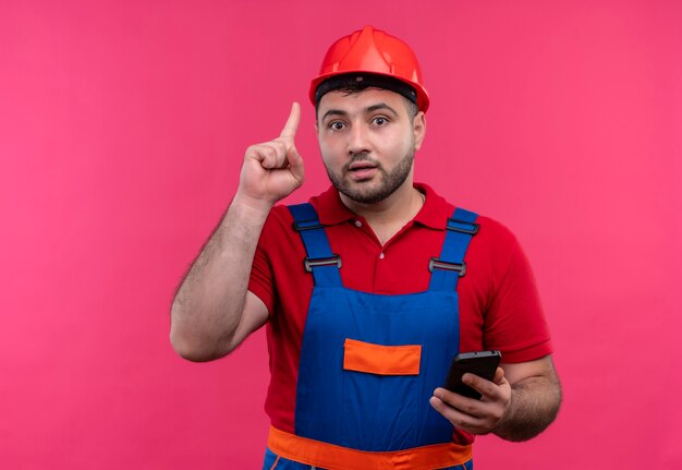 Молодой строитель в строительной форме и защитном шлеме держит смартфон, показывая указательный палец, напоминая себе о том, чтобы не забыть