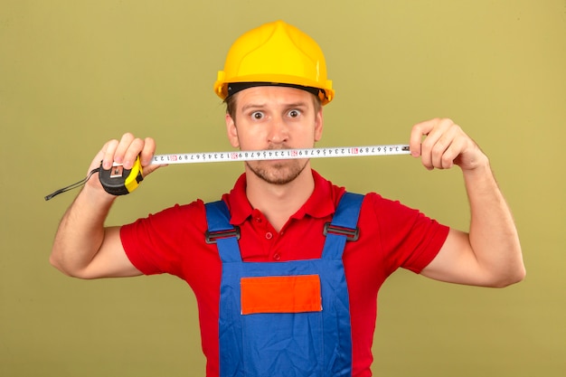 Young builder man in construction uniform and safety helmet holding tape-line with surprised face over isolated green wall