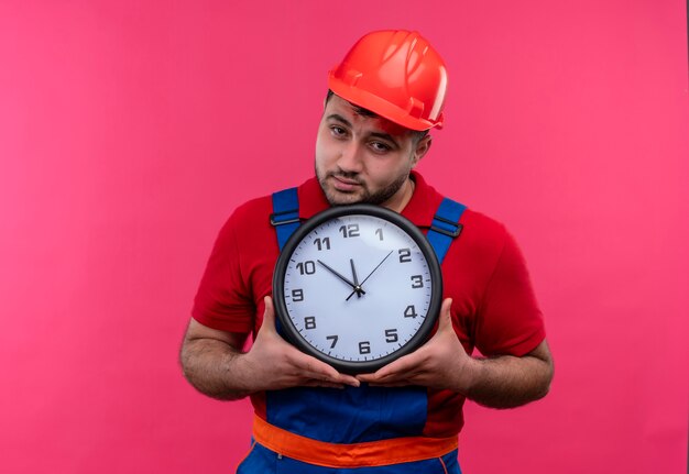 Young builder man in construction uniform and safety helmet holding wall clock looking at camera with unhappy face 