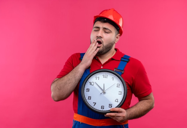 Young builder man in construction uniform and safety helmet holding wall clock yawning looking tired 