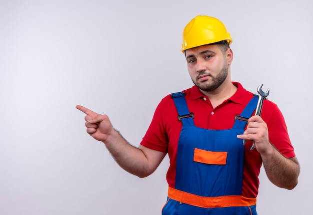 Young builder man in construction uniform and safety helmet holding wrench pointing with index finger to the side