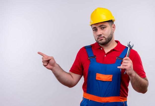 Young builder man in construction uniform and safety helmet holding wrench pointing with index finger to the side 
