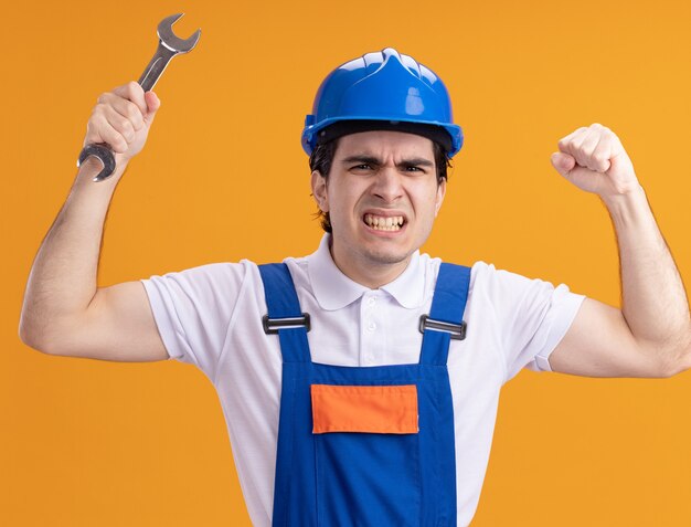 Young builder man in construction uniform and safety helmet holding wrench raising hands angry and displeased standing over orange wall