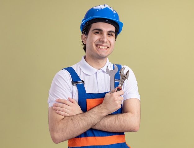 Young builder man in construction uniform and safety helmet holding wrenches looking at front with smile on face standing over green wall