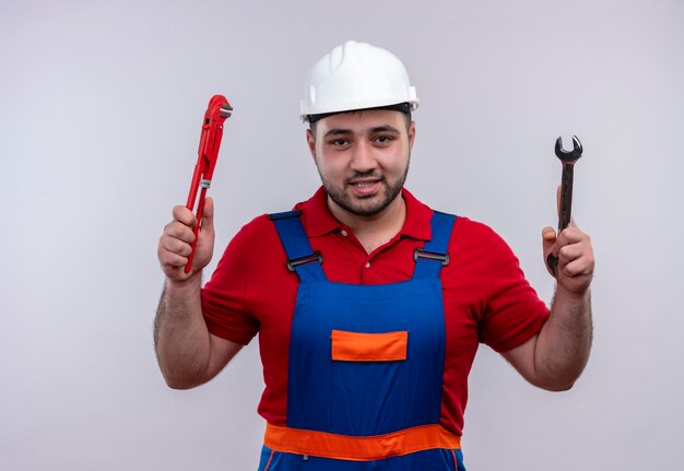 Young builder man in construction uniform and safety helmet holding wrenches in raised arms smiling 