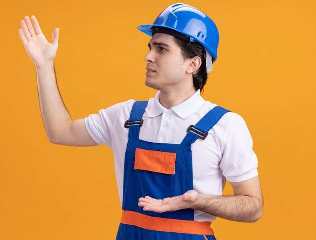 Young builder man in construction uniform and safety helmet looking aside being displeased gesturing with hands standing over orange wall