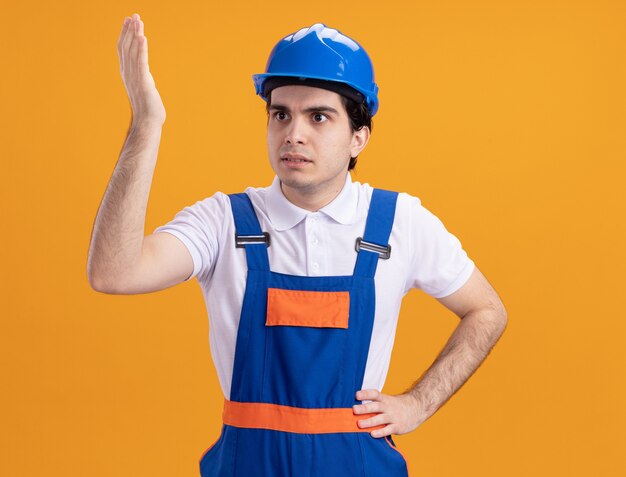 건설 유니폼과 안전 헬멧에 젊은 작성기 남자는 옆으로 오렌지 벽 위에 서있는 불만에 손을 제기 혼란 찾고