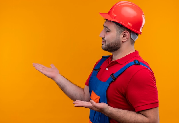 Young builder man in construction uniform and safety helmet looking aside presenting with arms oh hands 