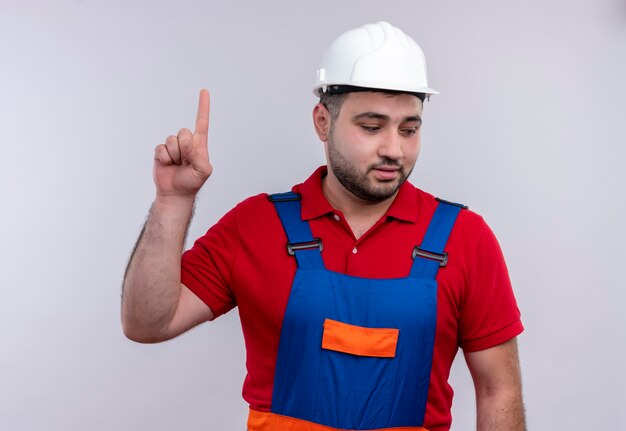 Young builder man in construction uniform and safety helmet looking aside smiling pointing with index finger up 