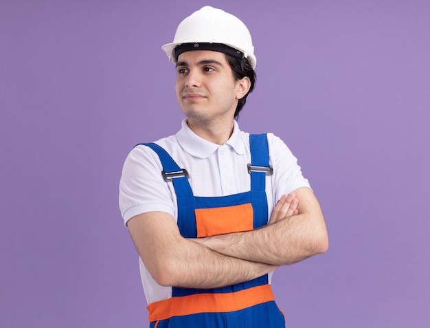 Молодой строитель в строительной форме и защитном шлеме, уверенно глядя в сторону со скрещенными руками, стоит над фиолетовой стеной