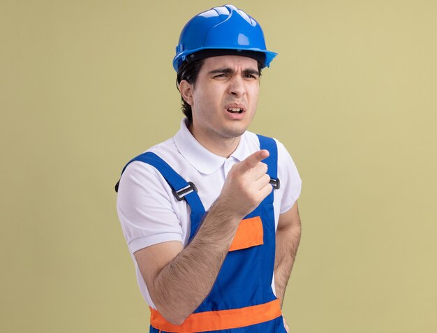 Молодой строитель в строительной форме и защитном шлеме смотрит в сторону с растерянным выражением лица, указывая указательным пальцем на что-то, стоящее над зеленой стеной