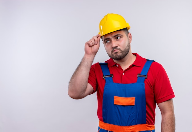 Молодой строитель в строительной форме и защитном шлеме смотрит в сторону со скептическим выражением лица и трогает шлем