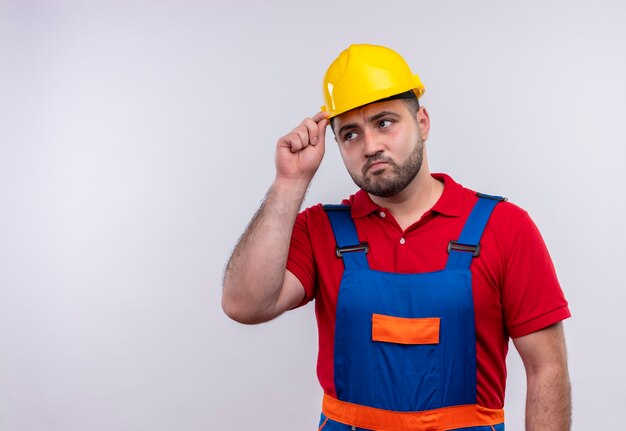 Young builder man in construction uniform and safety helmet looking aside with skeptic expression touching helmet 