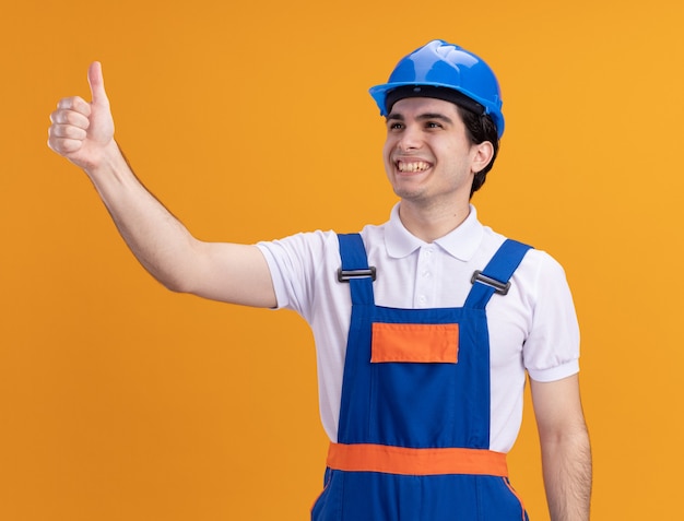 Young builder man in construction uniform and safety helmet looking aside with smile on face showing thumbs up standing over orange wall