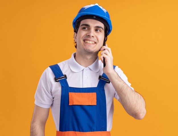 Young builder man in construction uniform and safety helmet looking aside with smile on face talking on mobile phone standing over orange wall