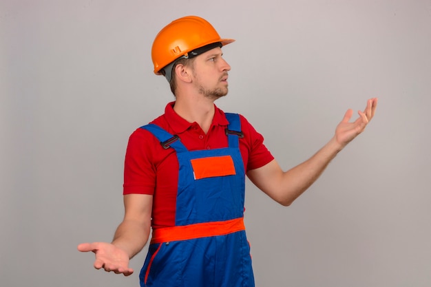 Young builder man in construction uniform and safety helmet looking away making confused gesture with hands and expression as asking question over isolated white wall