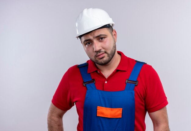 Молодой строитель в строительной форме и защитном шлеме смотрит в камеру со скептическим выражением лица