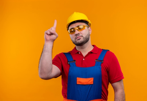 Foto gratuita uomo giovane costruttore in uniforme da costruzione e casco di sicurezza guardando fiducioso che punta con il dito indice su su sfondo arancione