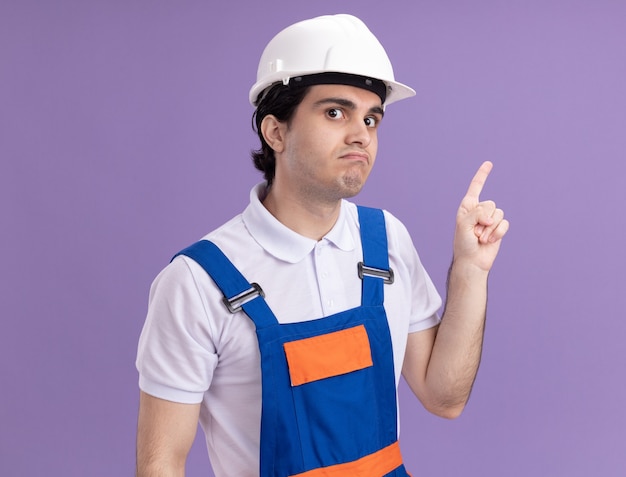 Молодой строитель в строительной форме и защитном шлеме выглядит смущенным, указывая указательным пальцем в сторону, стоя над фиолетовой стеной