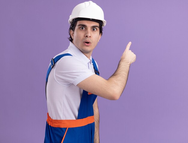 Young builder man in construction uniform and safety helmet looking confused pointing with index finger to the side standing over purple wall