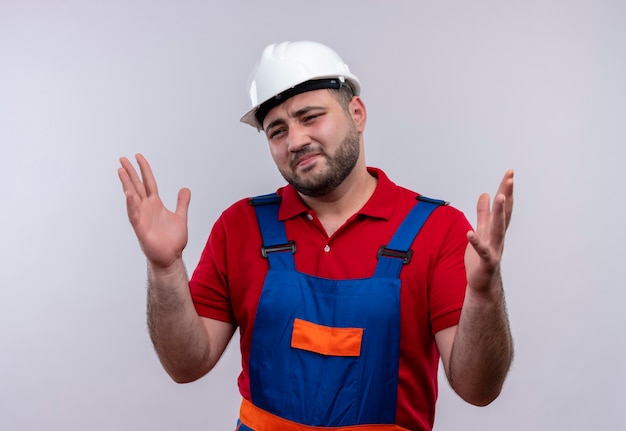 Young builder man in construction uniform and safety helmet  looking confused with annoyed expression   raising arms 