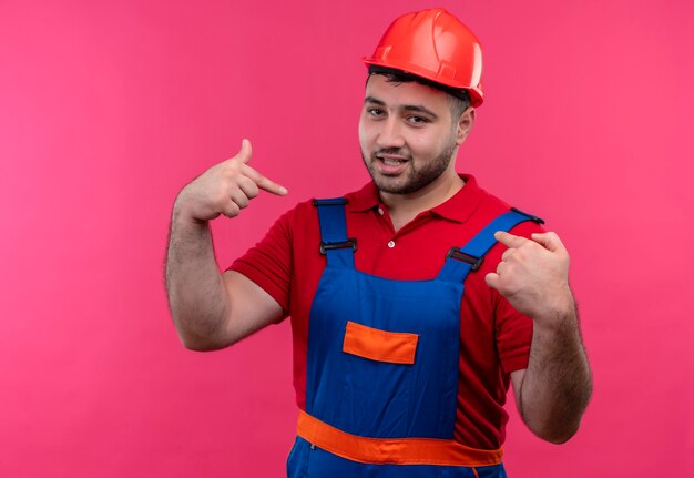 Young builder man in construction uniform and safety helmet looking feeling pround pointing with index fingers to himself 