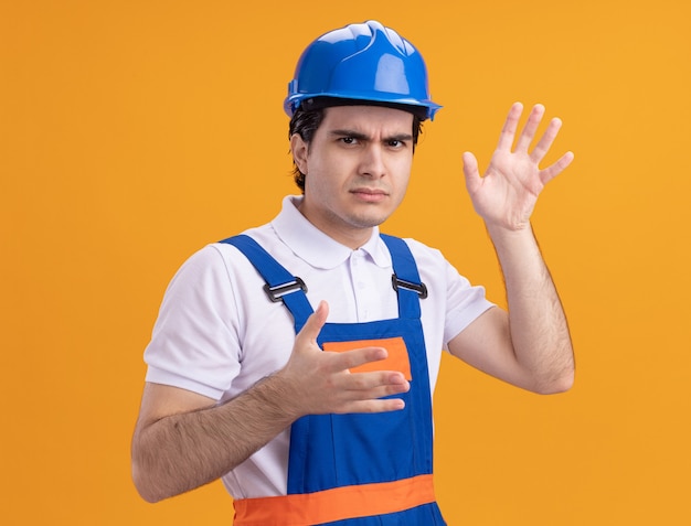 건설 유니폼과 안전 헬멧에 젊은 작성기 남자는 오렌지 벽 위에 서있는 팔로 불쾌 해하는 앞에서보고
