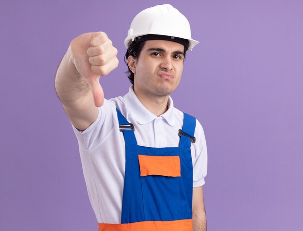 Молодой строитель в строительной форме и защитном шлеме, недовольно глядя на фронт, показывает большие пальцы вниз, стоя над фиолетовой стеной