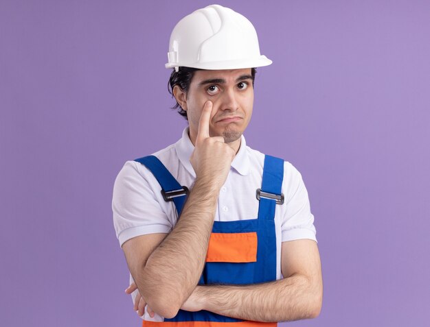 Young builder man in construction uniform and safety helmet looking at front pointing with index finger at his eye being confused standing over purple wall