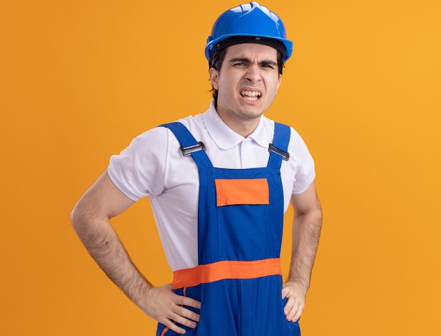 Foto gratuita uomo giovane costruttore in uniforme da costruzione e casco di sicurezza guardando la parte anteriore con la faccia arrabbiata delusa in piedi sopra la parete arancione