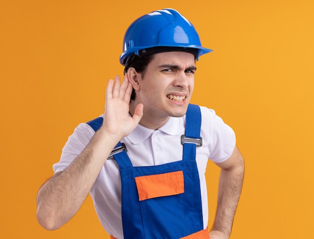 Foto gratuita uomo giovane costruttore in uniforme da costruzione e casco di sicurezza guardando la parte anteriore con la mano sull'orecchio cercando di ascoltare i pettegolezzi confusi in piedi sopra la parete arancione