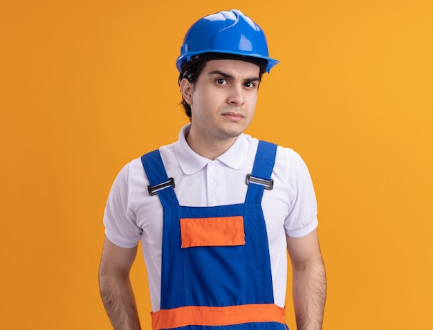 Foto gratuita uomo giovane costruttore in uniforme da costruzione e casco di sicurezza guardando la parte anteriore con espressione scettica in piedi sopra la parete arancione