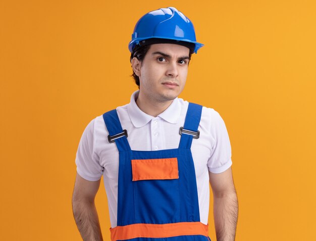 Young builder man in construction uniform and safety helmet looking at front with skeptic expression standing over orange wall