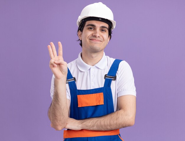 Young builder man in construction uniform and safety helmet looking at front with smile on face showing number three standing over purple wall