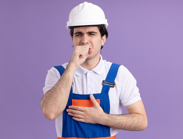 Young builder man in construction uniform and safety helmet looking unwell coughing standing over purple wall