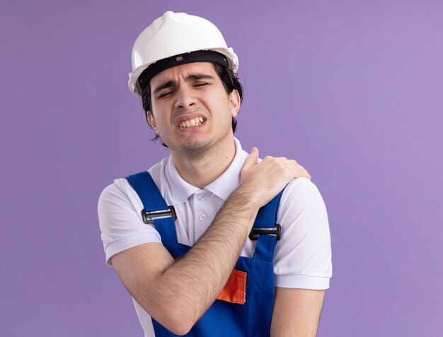 건설 유니폼과 안전 헬멧에 젊은 작성기 남자는 보라색 벽 위에 서있는 고통을 느끼는 그의 어깨를 만지고 몸이 좋지 않은 찾고