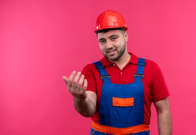 Молодой строитель в строительной униформе и защитном шлеме делает жест и дружелюбно улыбается рукой