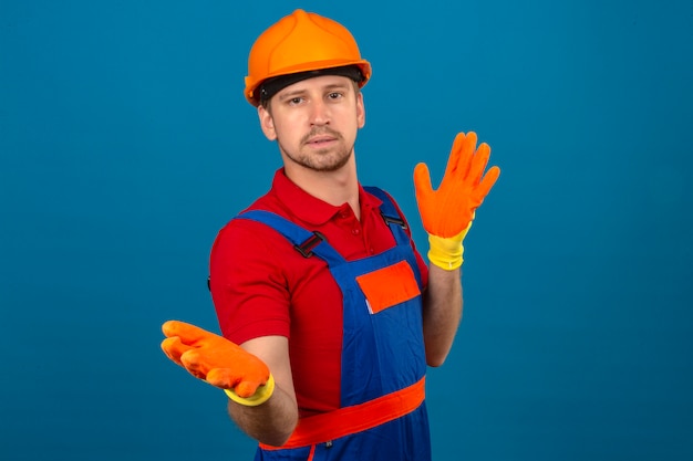 Молодой строитель человек в строительной форме и защитный шлем, делая путать жест с руками и выражением, как задавать вопрос на изолированной синей стене