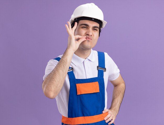 보라색 벽 위에 서있는 지퍼로 입을 닫는 것과 같은 손가락으로 침묵 제스처를 만드는 건설 유니폼 및 안전 헬멧에 젊은 작성기 남자