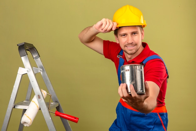 Young builder man in construction uniform and safety helmet on metal ladder holding paint can presenting to camera over isolated green wall