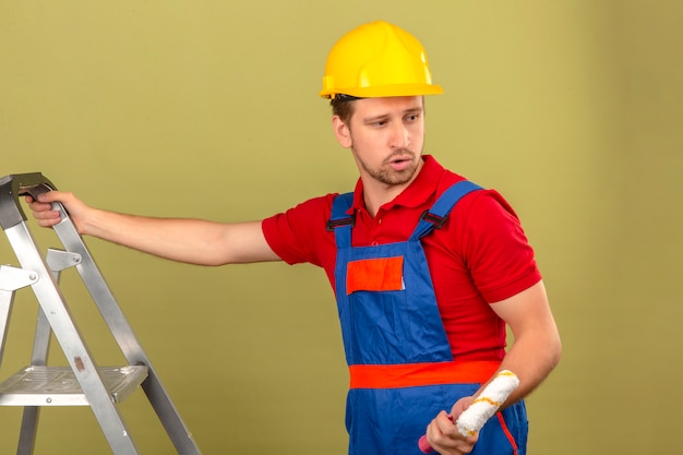 Young builder man in construction uniform and safety helmet on metal ladder holding paint roller looking sideways over isolated green wall