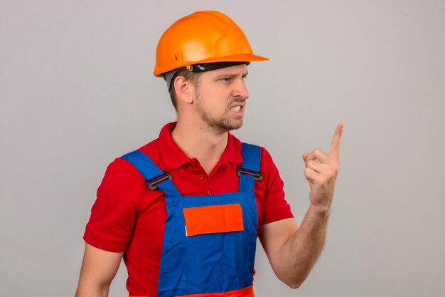 Young builder man in construction uniform and safety helmet pointing with finger up and angry expression over isolated white wall